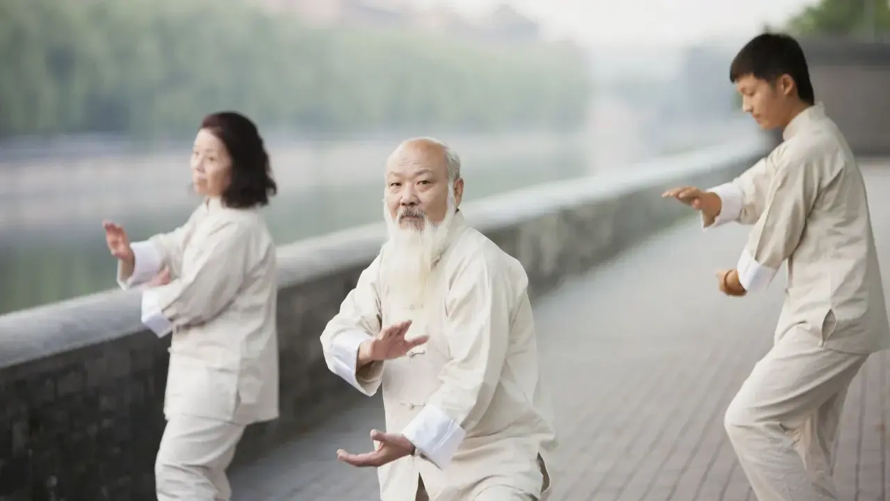 Trzy osoby ćwiczą tai chi nad rzeką. Starszy mężczyzna z białą brodą prowadzi grupę.