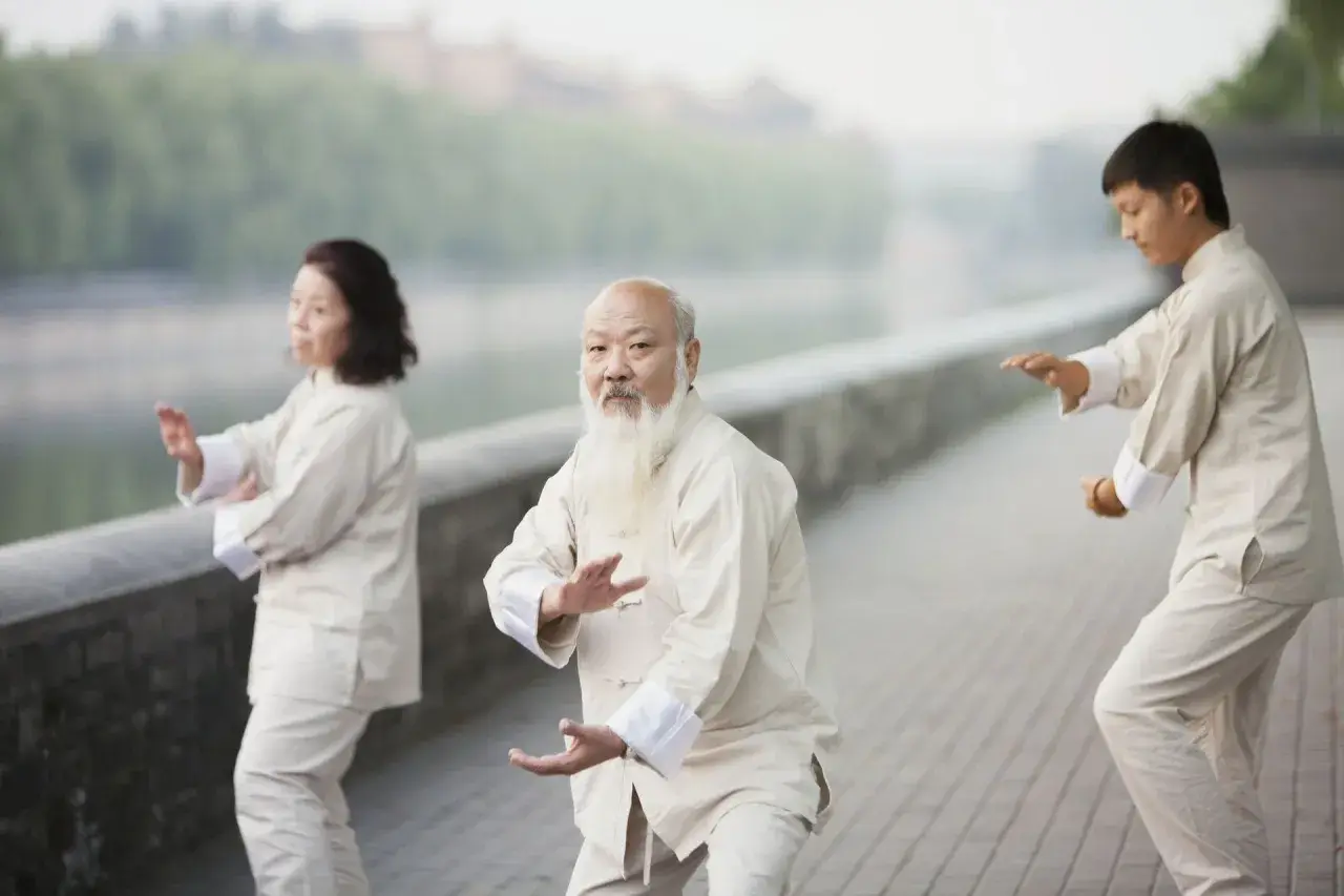 Trzy osoby ćwiczą tai chi nad rzeką. Starszy mężczyzna z białą brodą prowadzi grupę.
