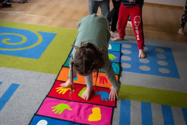 Dziecko wykonuje ćwiczenia na kolorowej macie z symbolami dłoni i stóp. Poduszka sensoryczna wspiera rozwój motoryczny.
